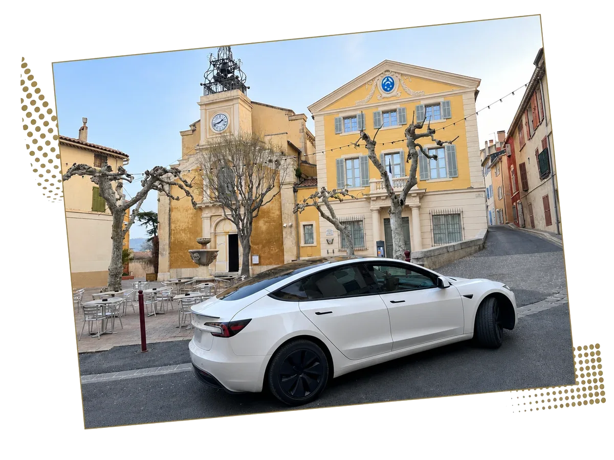 A white Tesla Model 3 in a square lined with Provençal facades